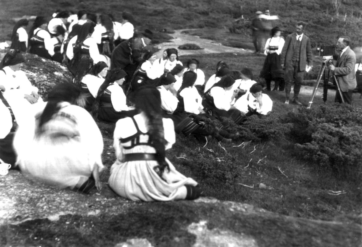 Kvinne- og mannsdrakt, gruppeportrett, Valle, Setesdal, Aust-Agder, antatt 1924. Gruppe sittende i terrenget utendørs med fotograf i aksjon til høyre. Fra "De Schreinerske samlinger" (skal oppgis).