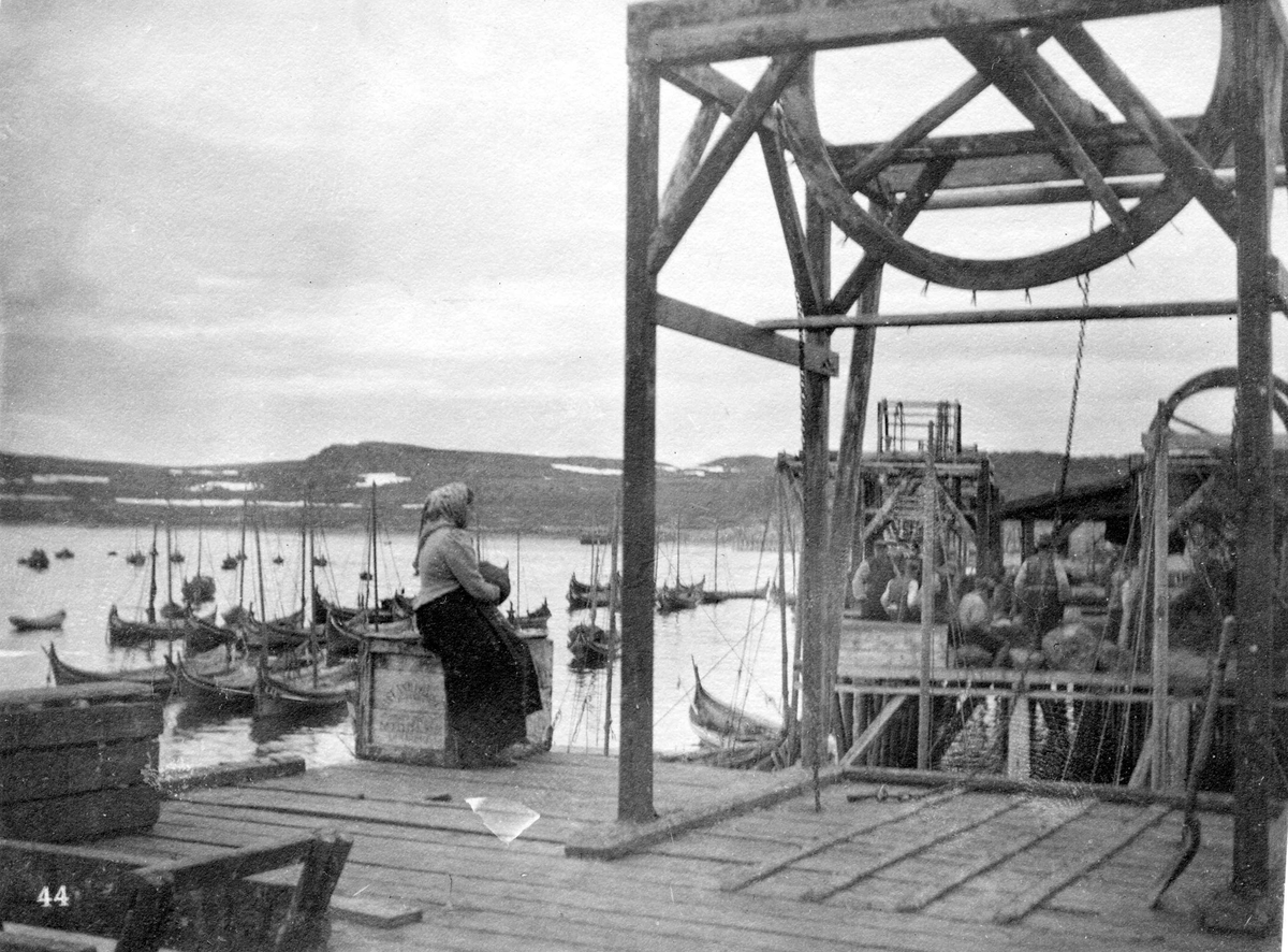 Kiberg fiskevær. Kvinne sittende på kasse på brygge. Fjord, Nordlandsbåter, fiskemottak.