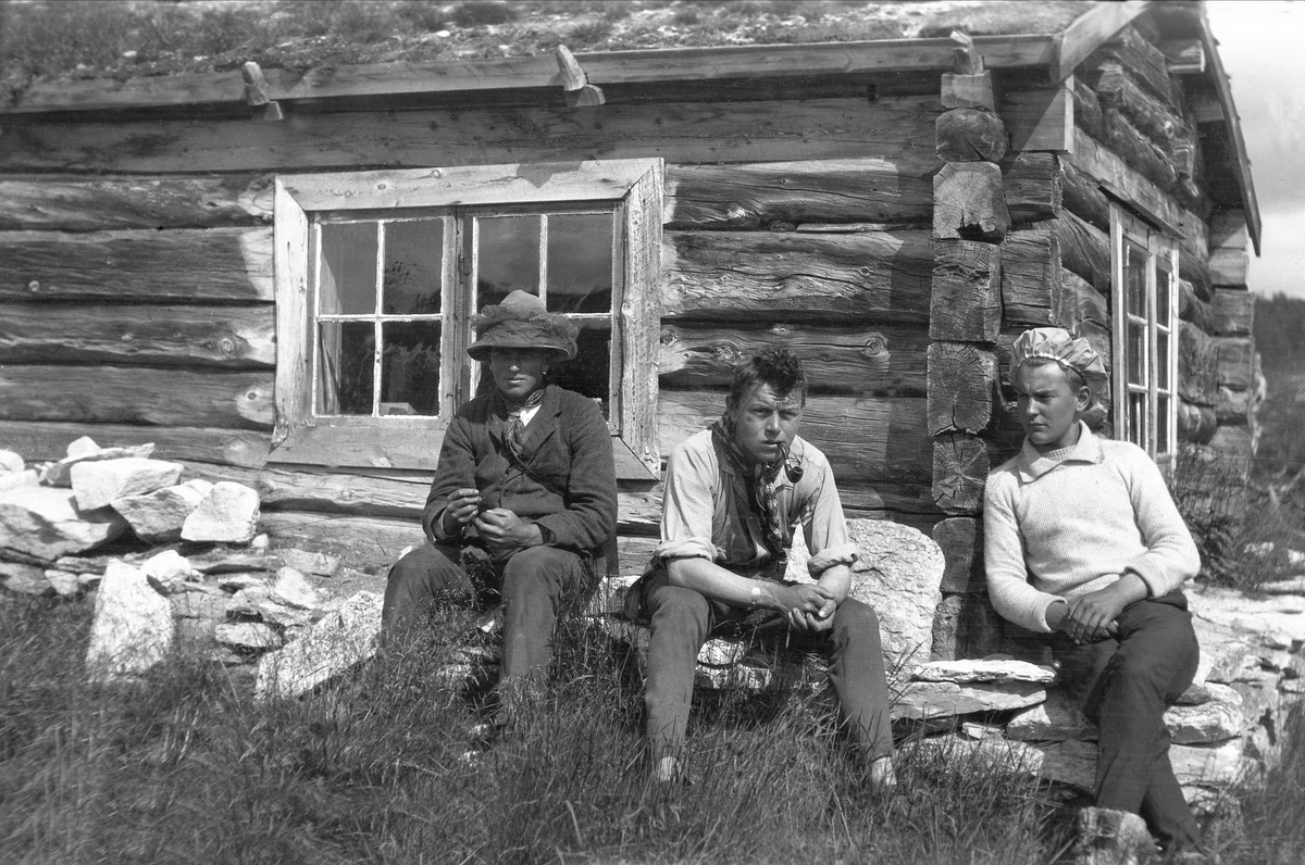 Tre menn sitter foran en tømmerhytte ved Mugga, Femundsmarka. Fotografert sommern 1922.