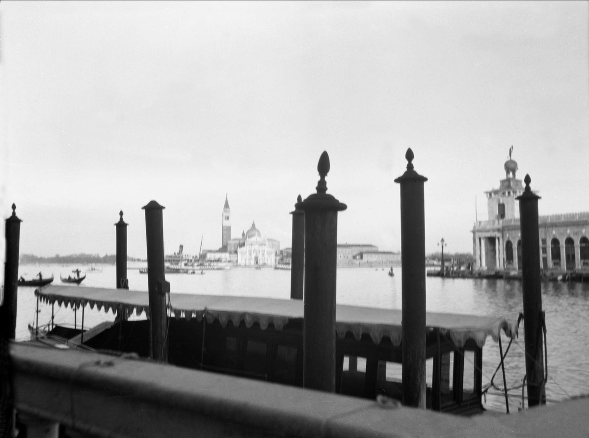 Bilde tatt fra en brygge, det viser noen forankringspåler for båter og gondoler. San Marco plassen er i bakgrunnen. Robsahm og Lund.