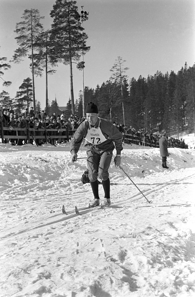 Skiløper med startnummer 72 i Holmenkollen. Publikum langs løypa.  Pressefotografer på sidelinjen. Holmenkollrennene 1963.