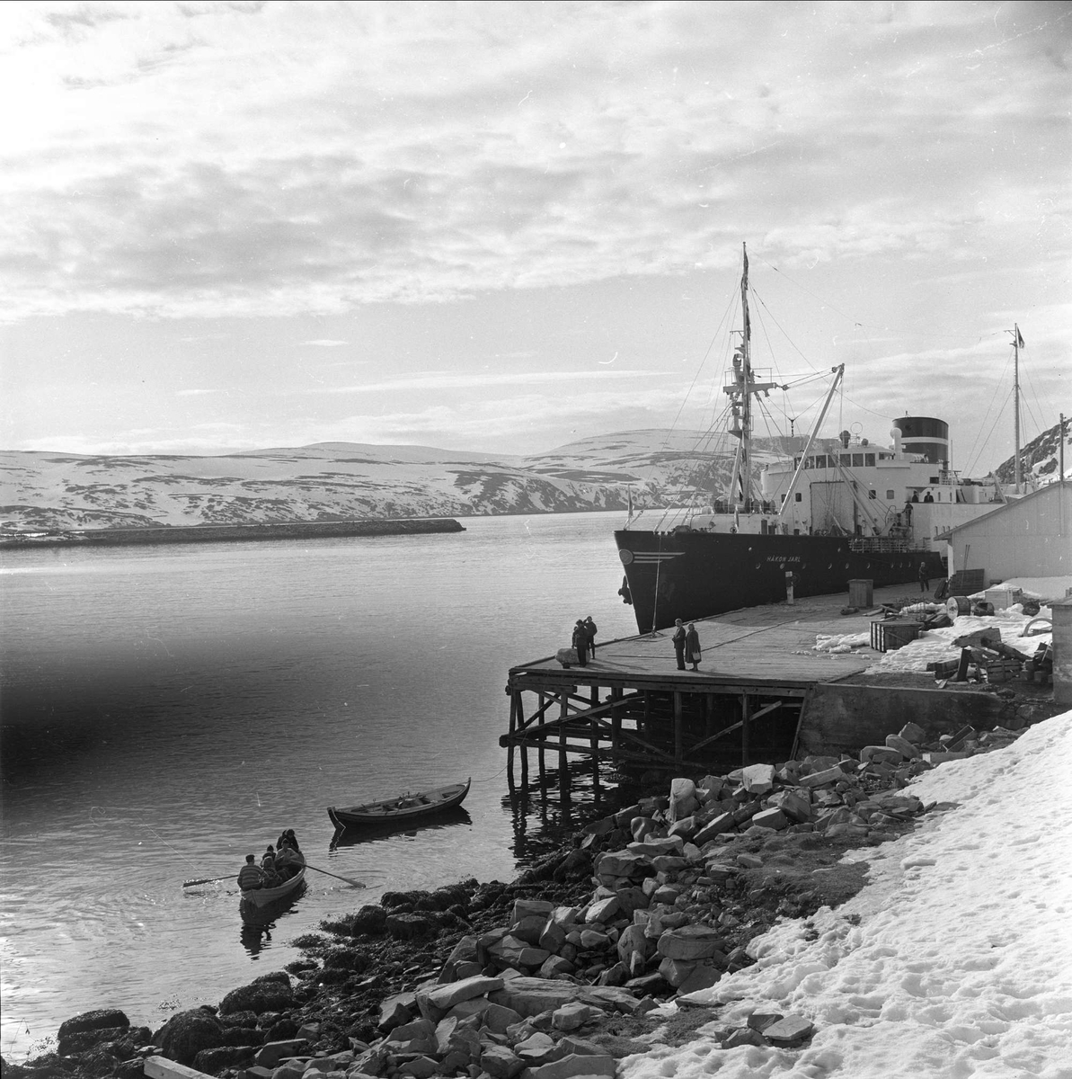Kjøllefjord, Lebesby, Finnmark, april 1963. Brygge med folk og båter. Hurtigruteskipet Håkon Jarl ligger ved kaia.