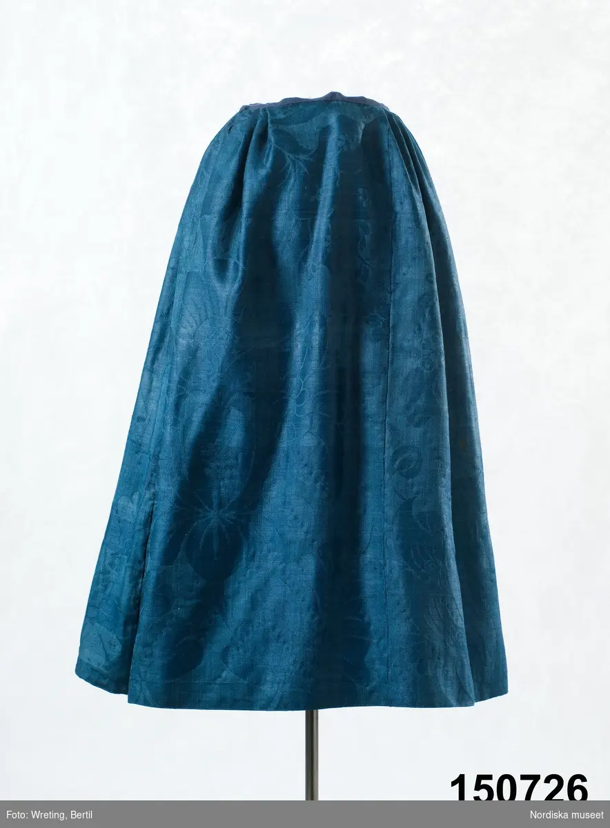Kjol av mörkblå ylledamast med ett mycket stort blommönster, 5 våder, varav en smalare. Vid ankomst till museet var kjolen uppsprättad till ett slätt tygstycke och har monterats till en kjol inför utställning i den modell som var vanlig i slutet av 1700-talet med slätt framparti 16 cm och därefter smala lagda veck gjorda på ett modernare sätt. Smal midjelinning av mörkblå linnelärft, sprund på höger sida. Skoning har sytts på av vit tapetserarväv.
Kjolen är blekt till en mellanblå färg, men den ursprungliga mörkt blå syns på avigan. Handsydd med blå lintråd.
Har enligt säljaren tillhört hans hustrus mormor, vars yngsta barn föddes 1844, och troddes därför kjolen kunna vara från 1820-talet. Troligen är den en generation äldre, eftersom dessa damasttyger slutade tillverkas runt 1800.
Dessa stormönstrade glättade ylledamasttyger var egentligen avsedda för inredning men blev under 1700-talets andra hälft använda  i bondemiljö som lyxplagg för rika bönder framförallt till bröllopsdräktens kjolar, västar eller livstycken. De finaste vävdes i Norwich i England, men kunde även vävas vid ylleväverier i Stockholm, dock inte lika  glättade.
/Berit Eldvik 2011-08-22