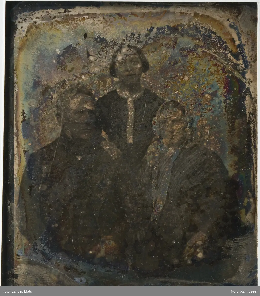 Grupporträtt av 3 personer, föreställer till höger, sittande, brandmästare Gustaf Wilhelm Draminsky, Landskrona och bredvid honom, till vänster, hans hustru Emilia Draminsky (född Hägg), längst bak, stående i mitten, deras fosterdotter, Emilia Fredrika Sophie (1828–1920). Dagerrotyp / daguerreotyp i ram. Plåten oxiderad, glaset sprucket, ramen i lösa delar. Baksida av mörkblå papp med små fastklistrade papperslappar med påskrift: "Mormor/Morfar/tant Emma". Nordiska museet inv.nr 198609
-
Portrait of Gustaf Wilhelm Draminsky, his wife Emilia and their foster daughter Emilia Fredrika Sophie. Oxidized sixth-plate daguerreotype in broken frame.