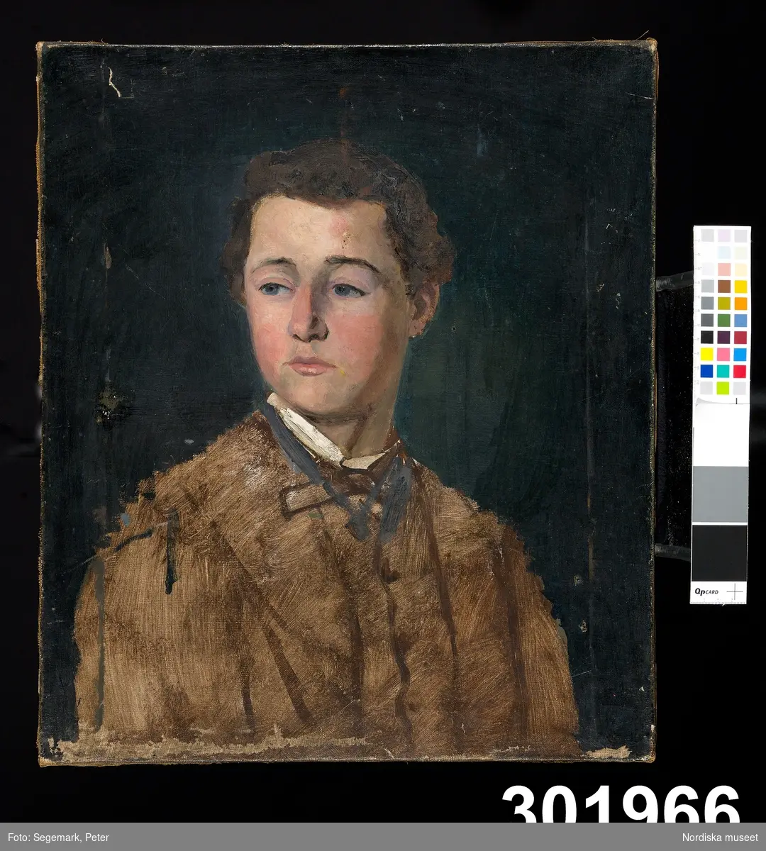 Porträtt, en face, av ung pojke i brun skjorta mot mörk bakgrund.