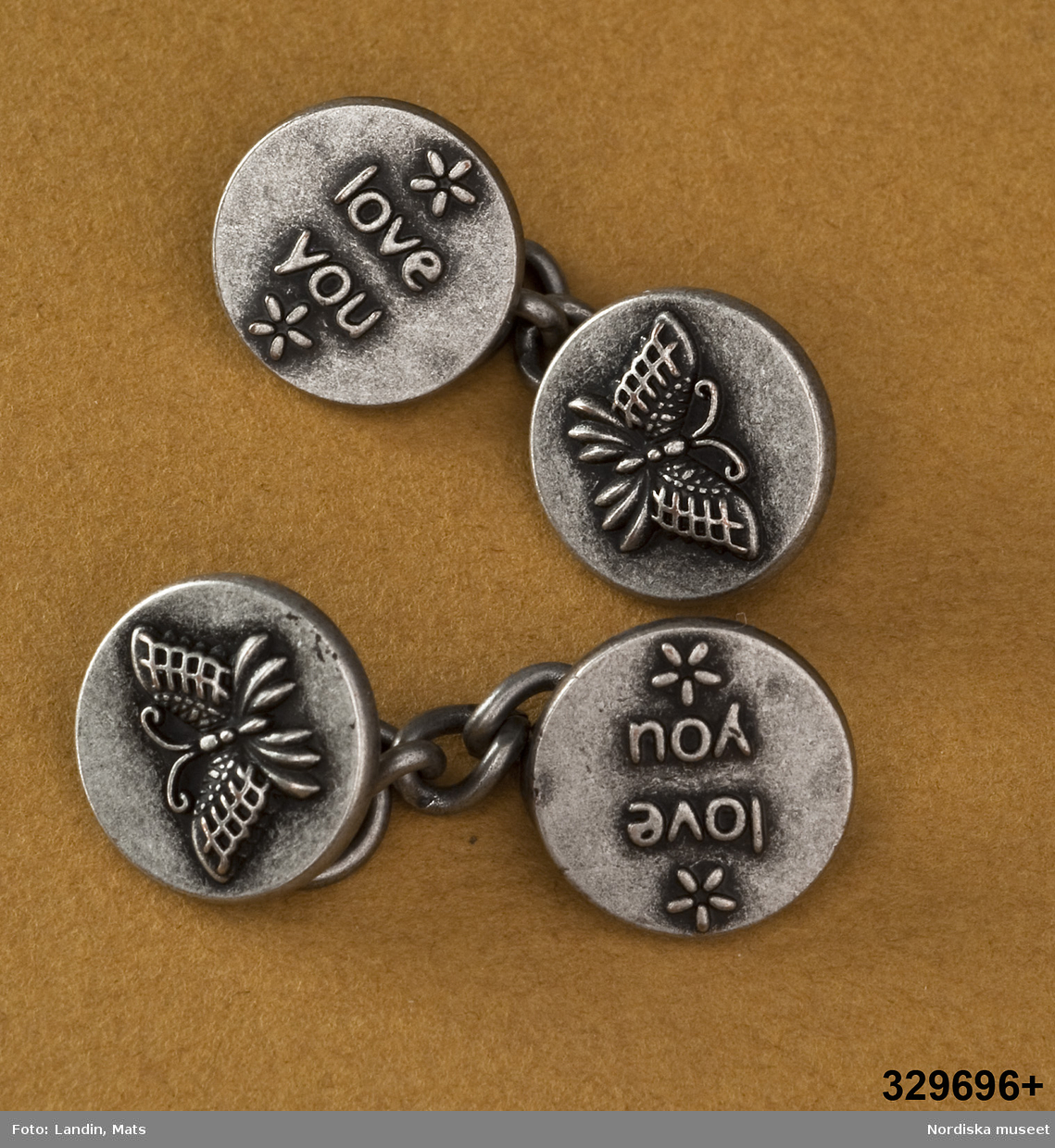 Ett par (2 st) länkknappar av vitmetall. bestående av två runda knappar med kedja emellan. På ena knappen en reliefbild i form av fjäril, på den andra text "love you". 
/Leif Wallin 2009-11-18