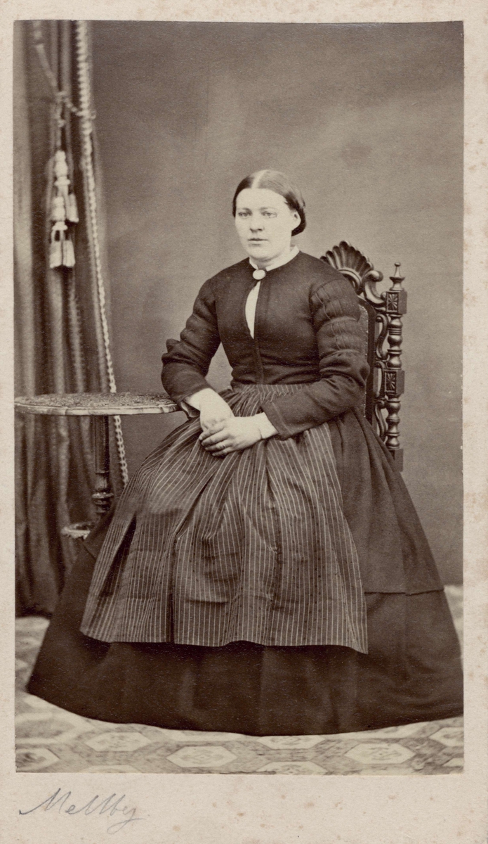 Porträtt av kvinna i en långärmad klänning och randigt förkläde sittande intill litet pelarbord. Mellby. Nordiska museet inv.nr. 18533A.