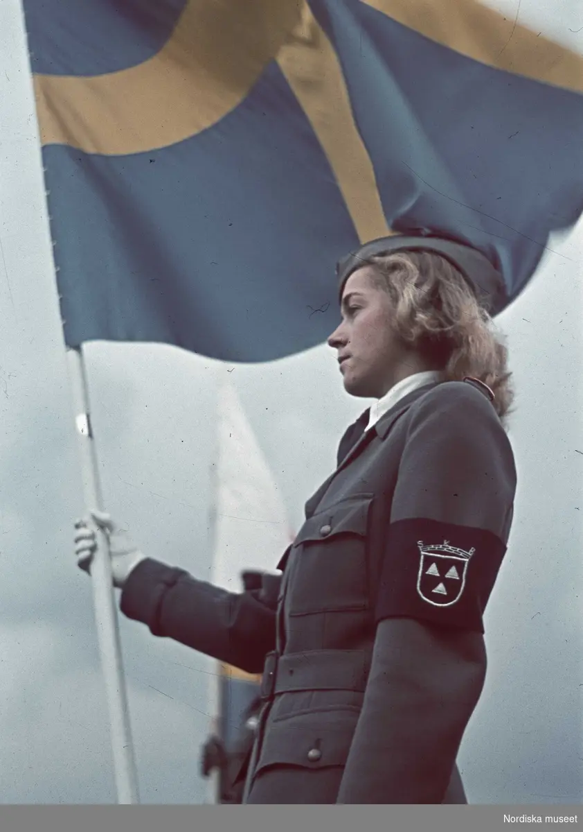 Porträtt av en ung kvinna i Sveriges Kvinnliga Bilkårers uniform. I ena handen håller hon en svensk fana.