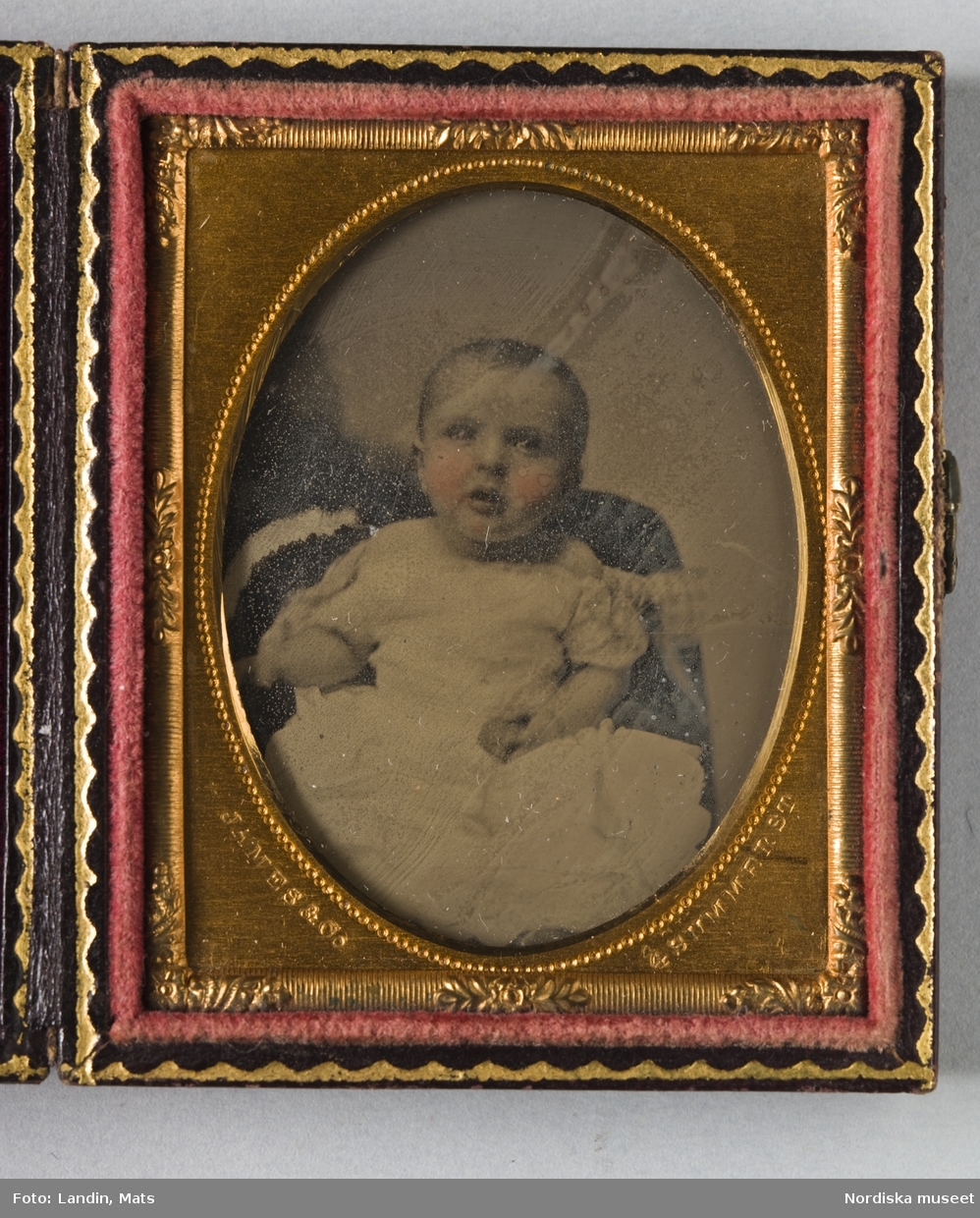 Porträtt av ett litet barn i vit klänning.Kolorerad Ambrotypi i förgylld metallram inpassad i fodral med sammetsfoder. Instansat i passepartouten: "James & Co, Summer St, 1860."
Nordiska museet inv.nr 279402
