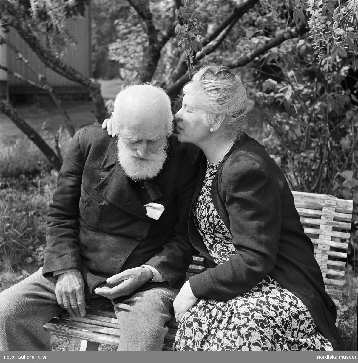 Den norske författaren Knut Hamsun tillsammans med makan Marie vid parets bostad i Norge i maj 1950. Paret sitter på en bänk i trädgården. Hon böjer sig fram mot honom, kanske för att viska något i hans öra.