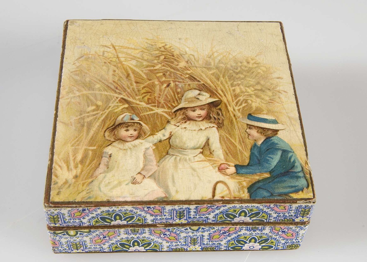 Dekor i blått, gult, rosa och vitt. På locket bild av tre barn. På lockets insida vit etikett med svart text: Th. Hagmann, Guldsmed, Upsala.