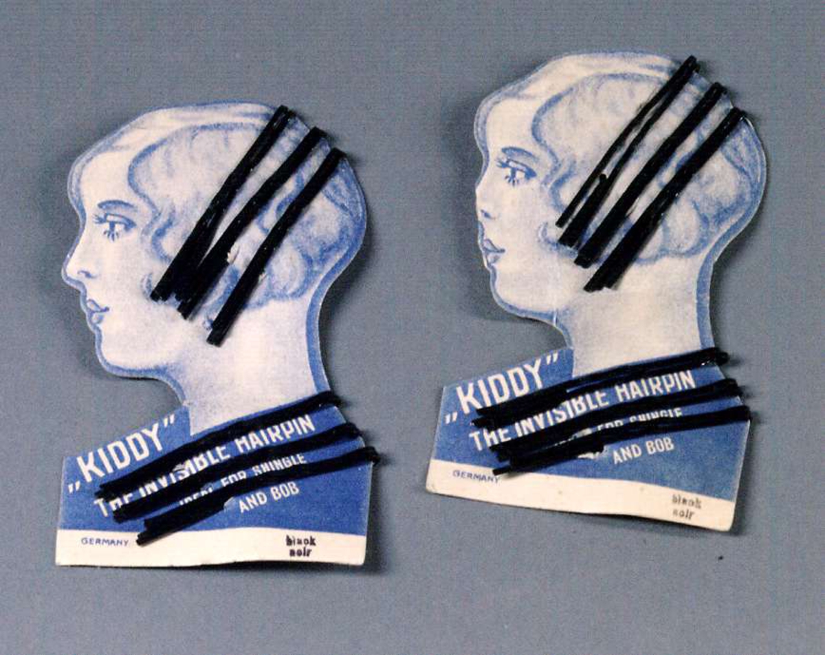 Två förpackningar med 12 smala hårspännen av metall. Förpackningen i blå papp formad som kvinnoansikte, vit text: "KIDDY" THE INVISIBLE HAIRPIN.