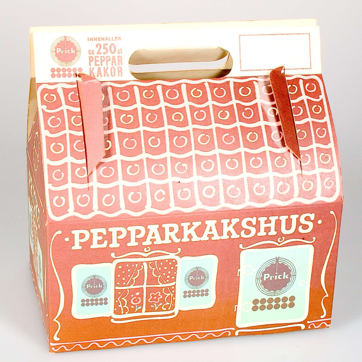 Pepparkaksförpackning av papp, utformad som ett pepparkakshus. Tryckt dekor i brunt, rosa och blått. Logotypen Prick på varje sida av förpackningen samt texten: PRICKS PEPPARKAKSHUS, INNEHÅLLER ca 250 PEPPARKAKOR.
