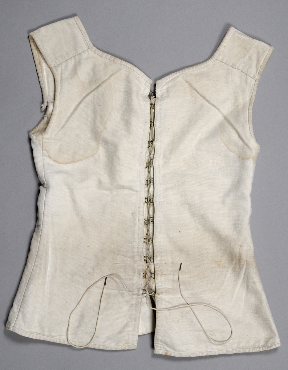 Snörliv, av vit bomull (kypert). Helt handsytt. Två delar som snörs samman bak. Snöret löper genom metallhyskor. Knäpps framtill med sex knappar (fem porslin och en metall). 1840-60-tal (nyrokoko).