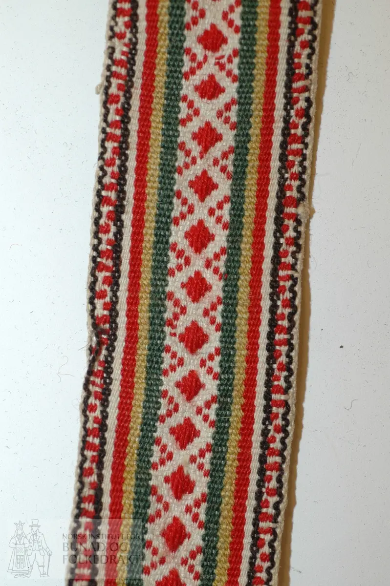 Band, innslagstråd av lin, og renningstråd av kvit bomull og ulltråd i fargane brun/svart, raudt, grønt og gult.