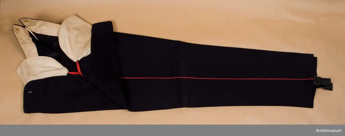 Ylle. Storlek: B 252. Mörkblå långbyxor med röd passpoal i sidsömmen.  Försedda med resår nedtill som knäppes fast på innersidan av  sidsömmarna. Resåren tjänar att sträcka byxan och skall sitta  under skodonets hålfot.