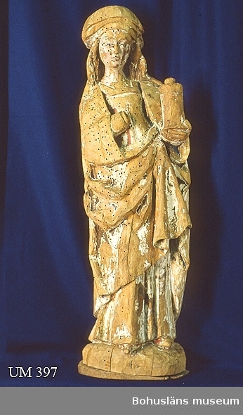 Träskulptur från 1500-talets början. Skulpturen bör ha tillhört ett medeltida altarskåp, möjligen tillsammans med skulptur UM000396 och UM000398.
Skulpturen håller ett smörjelsekärl eller en salvburk i handen vilket brukar vara Maria Magdalenas helgonattribut.
Hela skulpturen har varit bemålad, polykrom träskulptur. Träkärna utskuren i lind i ett helt stycke så när som på högerhand, vilken tillverkats separat och satts fast. Handen saknas. Rester av linneduk finns i plugghålet. Även i huvudet finns rester, även träplugg och linneduk. En bottenplatta ät fastsatt med två träpluggar och en spik, som tillkommit senare.
Skulpturen har varit bemålad och har haft en lim - krita grundering över hela ytan, på vissa ställen ca. 1 mm tjock. Rester av linneduk finns på högersida av manteln och framtill på klänningen finns tyg och trådrester för att dölja ett kvisthål i träet. 
Karnationen är målad i oljefärg. Av denna finns spår av rosa kinder, läppar och hals. Ögonen har svarta konturlinjer. Håret har rester av brun-svart färg och bronsering. På klänningen finns röd färg och rester av en mönsterbård med gröna pigment. På manteln finns rester av brunt, rött och blått laserade på varandra. I nederkanten av manteln finns grön färg och fördjupningar av streck dragna genom färg och grundering. Guldglans kan skönjas på vänstersida av manteln. Insidan av manteln har varit blå, målad med ultramarin, lapis lazuli. Klädedräkten är målad med temperafärg.

Ur handskrivna katalogen 1957-1958:
S:ta Sunniva, Krokstad
H. 82 cm; i vänster hand smörjelsekärl, höger avslagen; färgen flagn. maskäten. 
Maria Magdalena.