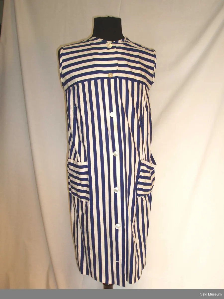 Ermeløs kjole i hvit/blå stripete stoff. Knepping i hele kjolens lengde. Stoffbelte i samme mønster.