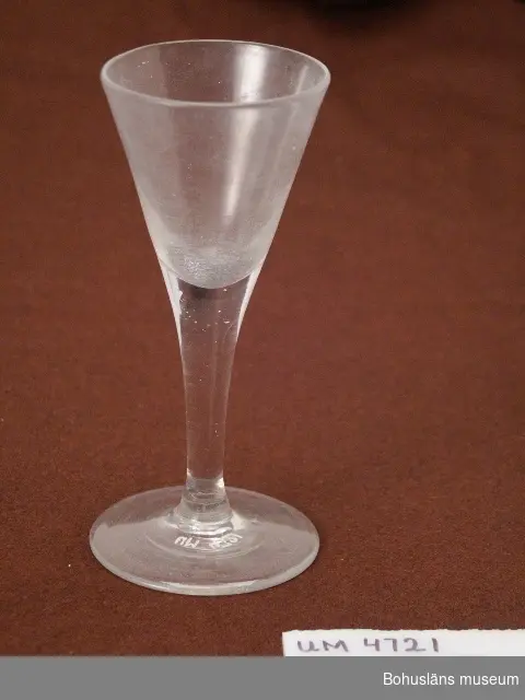 Spetsglas för vin och brännvin. Stor cuppa, fotring med lite större diameter än cuppan. Puntelmärke.
Glaset har en mängd smårepor på insidan vilket ger det ett gråaktigt utseende.

Ur handskrivna katalogen 1957-1958:
Brännvinsglas 16 cm.
Mynningsdiamet.: 7 cm. Fotens diamet.: 7,6 cm. Vitt glas, slätt. Föremålet helt.

Lappkatalog: 61

Litt: Hermelin, C. F., Welander, E.: Glasboken. Historia, teknik och form. Askild & Kärnekull 1980.
Nisbeth, Å. Glas i Sverige intill 1900. Ur Svenskt glas. Wahlström & Widstrand 1991.
Steenberg, E.: Flaskor och glas. Nordiska museet 1952.
Steenberg, E.: Svenskt glas. Bohusläningens AB Uddevalla 1964.