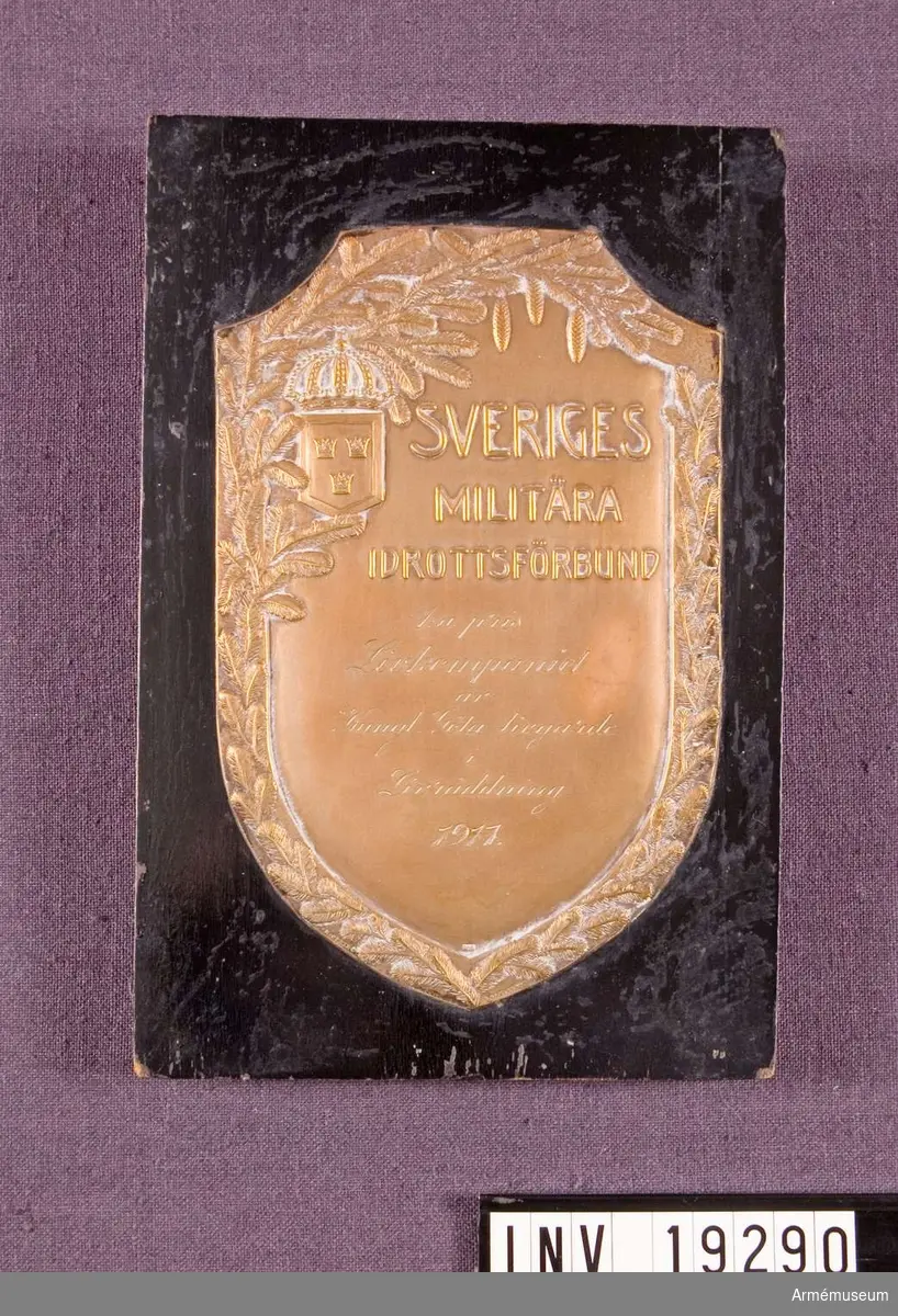 Plakett i brons med text: "SVERIGES MILITÄRA IDROTTSFÖRBUND 1:a pris Livkompaniet av Kungl. Göta Livgarde i Livräddning 1917." Plaketten fästad på svart lackad träplatta.