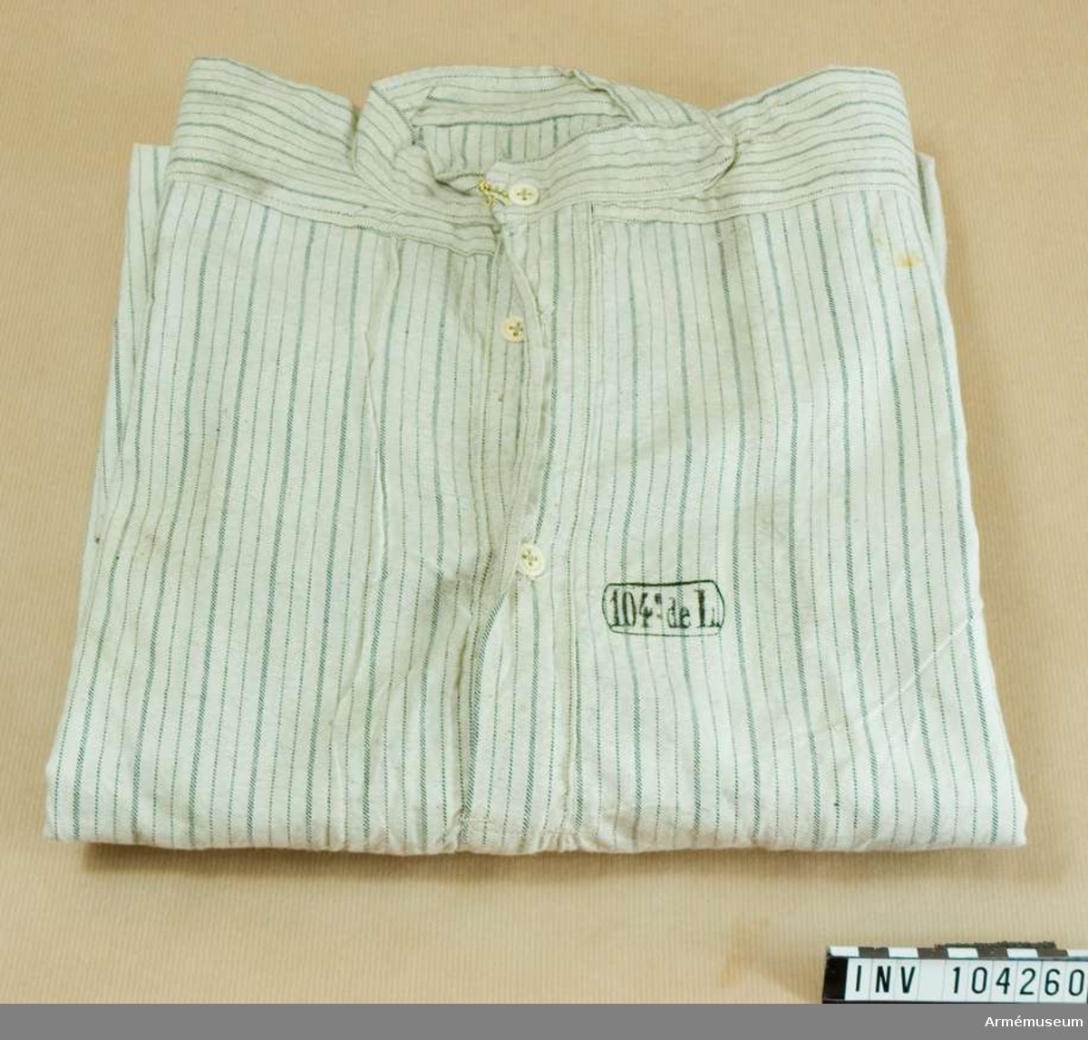 Skjorta av bomullstyg (vit med gråa ränder) med fast upprättstående krage och långa ärmar. På bröstets öppning två knappar, och på krage en; alla äro små, vita av ben. Likadana knappar, en å varje, på ärmuppslagens sprund. På skjortans framsida, en stämpel: "104de L" - 104 linjeinfanteriregementet. På skjortans baksida stämpel "30.11. 1910" - allt annat oläsligt.