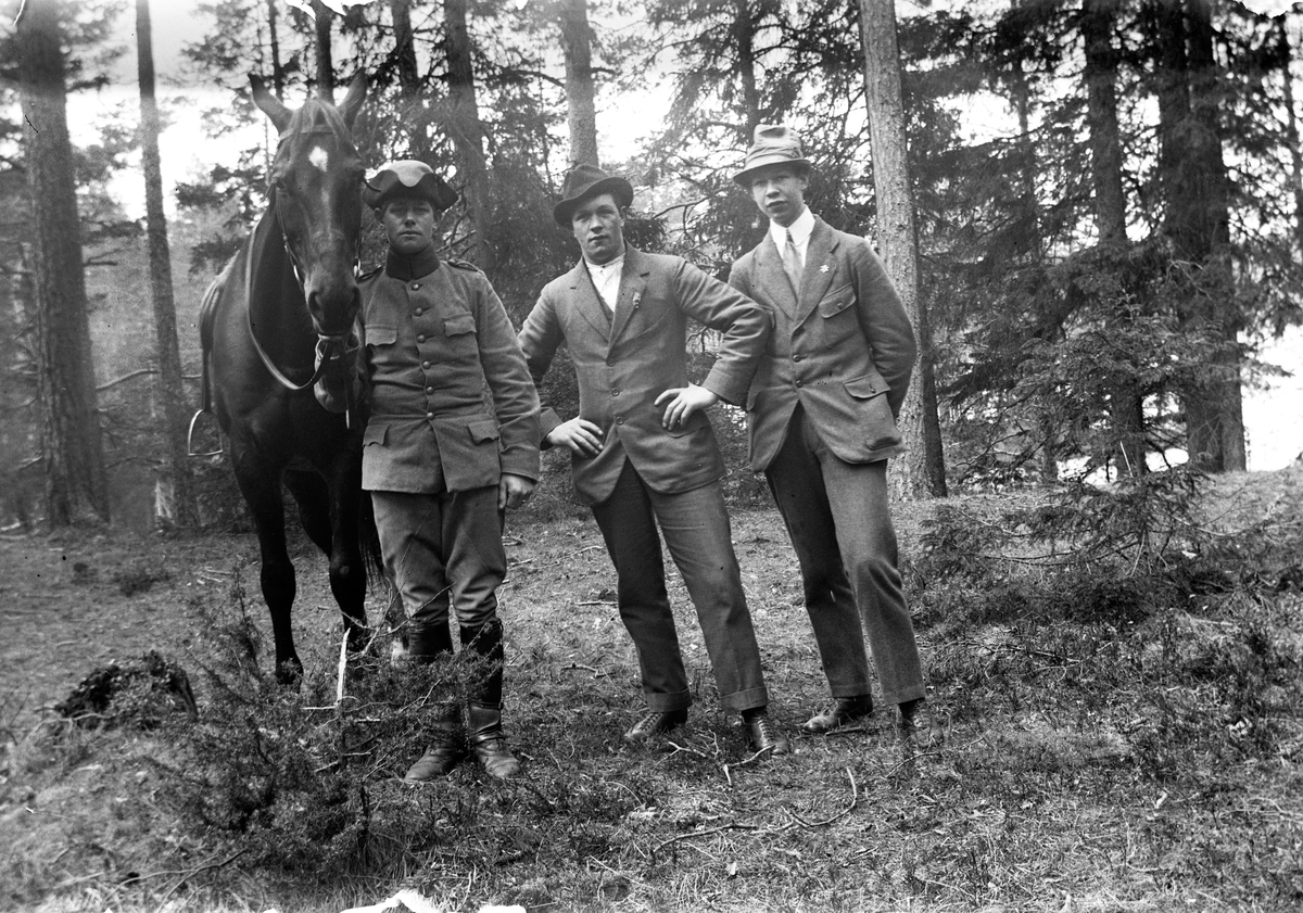 Tre män varav en i soldatuniform, samt en ridhäst i barrskogen.