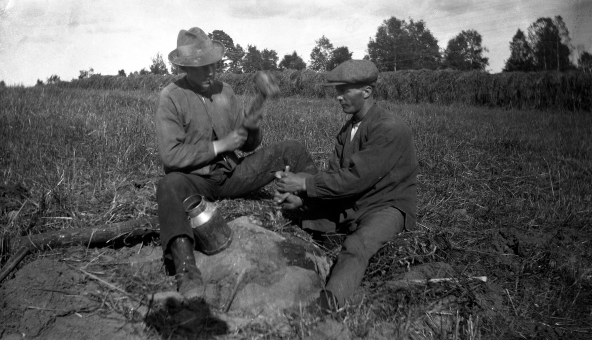 Två män försöker spräcka en sten på en åker.