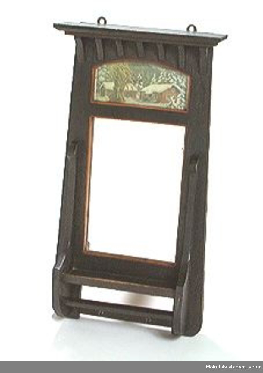 Svartmålad spegel med en hylla ovanför spegelglaset och en nedanför. Ovanför spegelglaset finns bakom glas två röda stugor i ett vinterlandskap jämte en julkärve med en bofink (Allersmotiv från 1920-talet).