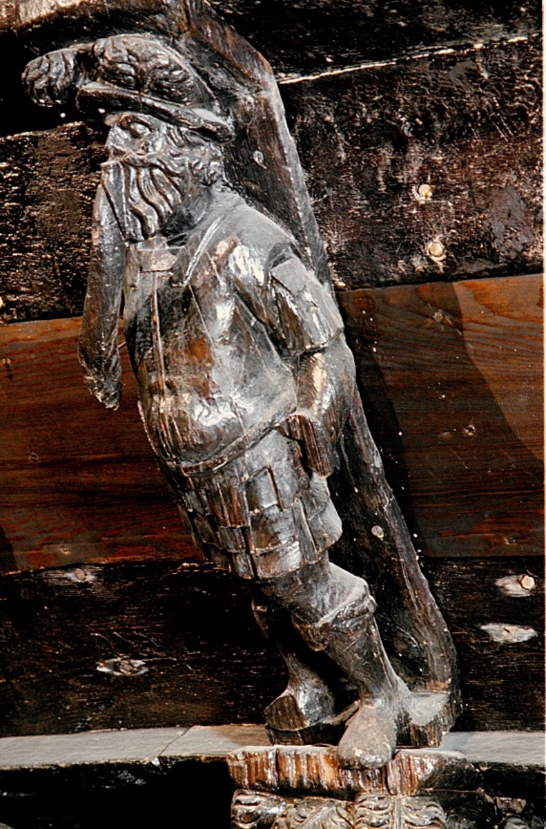 Krigare iklädd romersk rustning och en bredbrättad plymhatt. Mannen har långt, lockigt skägg och mustascher. Huvudet är något vridet åt höger. Rustningens nederdel består av ett undre lager stora, rektangulära och metallskodda flikar över tunika samt ett övre lager mindre flikar av samma form, omväxlande med halvrunda flikar. Mannens mage är kraftigt markerad. De separat snidade ytterdelarna till armar och händer saknas. Ett mindre parti av vänster arm har dock påträffats löst. Delar av fingrar till höger hand kan skönjas, hållande ett stort, något svängt instrument - tuba eller dylikt - i vilket mannen tycks blåsa. Instrumentet är snidat i ett med bålen. I vänster hand har mannen troligtvis hållit en fackla eller lampa. Kroppstyngden vilar på höger ben medan vänster ben är något böjt. Främre delen av höger fot har fallit bort. Vänster fot sticker ut ett bra stycke utanför den minimala ståplattan.
Bortsett från ovannämnda defekter samt ett visst slitage är skulpturen relativt välbevarad.

Text in English: Warrior clad in Roman armour, with long curly beard and long thin moustache, wearing wide-brimmed helmet or hat. The head is slightly turned to the right. The stomach or belly of the figure is well defined. The ends of the arms and hands are missing. A smaller section of the left arm was found separately. Parts of the fingers on the right hand can be seen holding a large spiralled instrument being blown by the figure. The instrument is carved from the same piece of wood as the trunk of the figure. The figure should have been holding a torch or lamp in his left hand. The weight of the body rests on the right foot while the left leg leg is slightly bent. The toe section of the right foot has fallen off. The left foot protrudes quite a distance over the small foot base.
Aside from the above-mentioned defects and a certain degree of wear, the sculpture is rather well preserved.