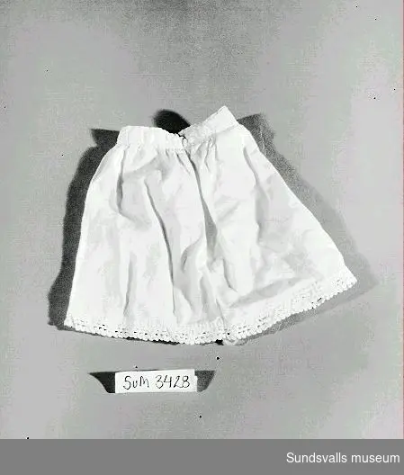Underkjol för docka av vitt bomullstyg med vit spets i nederkanten. Knäppes med plastknapp bak.