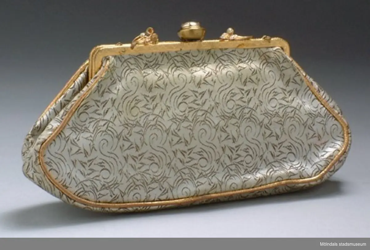 Liten oval handväska med "portmonnä" öppning. Den är vit och är guld/brun mönstrad. Öppningen är guldfärgad och har stenar som utsmyckning. Har troligtvis haft ett axelband, men är borta.Måtten:Bredd 220 mm, höjd 120 mm.
