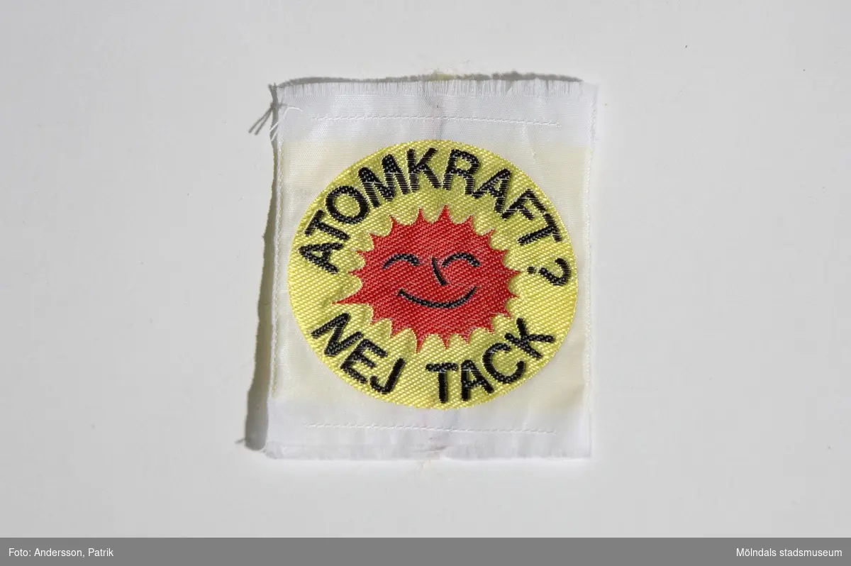 Tygmärke med symbolen och texten: "ATOMKRAFT? NEJ TACK" Märket är sytt på en vit fyrkantig tygbit. Märket är runt med gul bakgrund och har den orange symbolen med texten i svart.Detta användes av Lena Måvholm 1979 i samband med  folkomröstningen om kärnkraft i Sverige som skedde 1980.MåttLängd: 51 mm, Bredd: ca. 61 mm, Diam: ca. 47 mm.1980 skedde en folkomröstning om kärnkraften i Sverige. Då var det många personersom under 1970 och 80-talet bar på denna symbol. Antikärnkraftsymbolen liknade ett glatt rött/orange eldklot och hade texten: â€ATOMKRAFT? NEJ TACKâ€. Det var en livskraftig symbol som ville visa energi och framtidstro. Den ville visa vad man borde använt sig av i stället såsom solen, naturen och vinden.