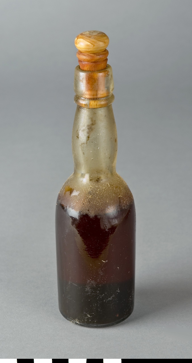 Flaska av ofärgat glas med okänd grön vätska. En svaravad kork av trä försluter flaskan.