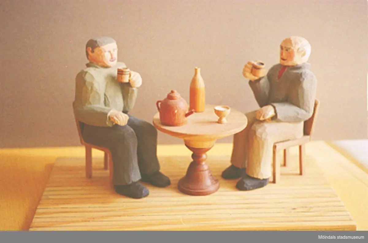 Två gubbar sitter på var sin stol vid ett runt bord och dricker ur varsitt ölkrus. På bordet står en kaffekittel, en sockerskål och en vinflaska. Harry Bergmans "gubbar" (träfigurer). Liknande foto 1994_0965.