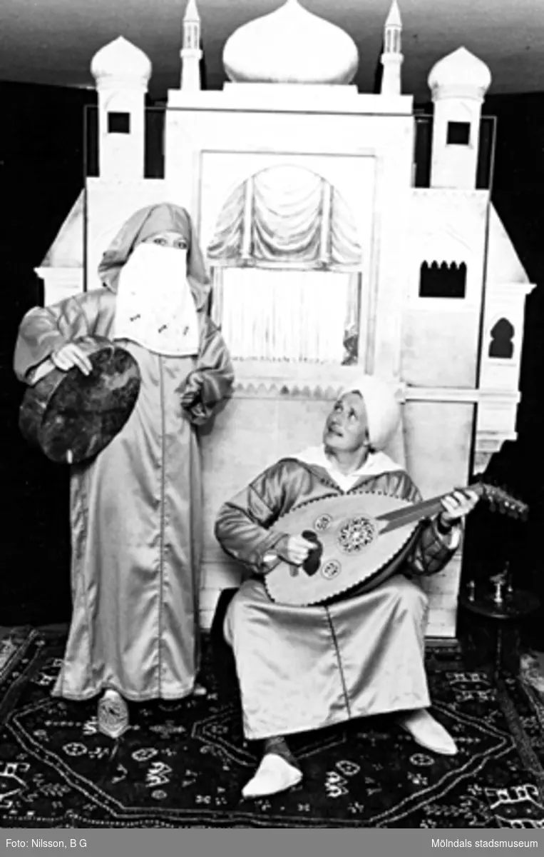 Två vuxna personer utklädda i orientaliska kläder, båda hållandes var sitt instrument. I bakgrunden står ett orientaliskt palats i mini-format. Holtermanska daghemmet 1973.