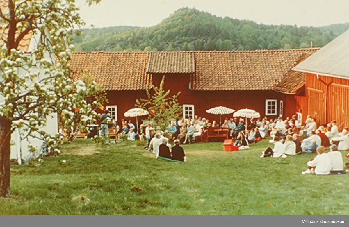 Människor sitter i gräset och på bänkar utanför Börjesgårdeen/Lindome Hembygdsgård. De lyssnar på ett uppträdande. Okänt årtal.