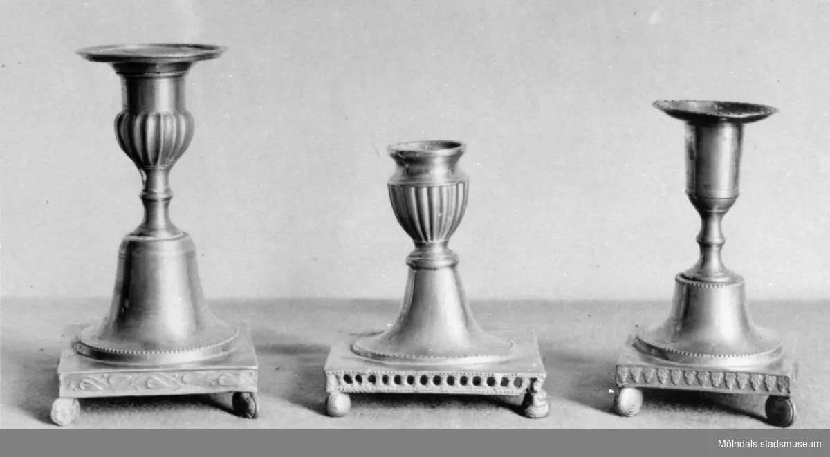 Tre ljusstakar av metall med varierande design och höjd. Samtliga står på var sin sockel. Gunnebo slott, 1930-tal.