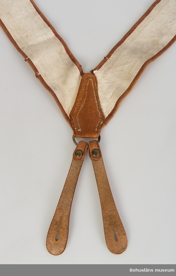 Ett par hängslen. Broderade, troligen tillverkade under 1840-1850-talet. Randig bottenväv med korsstygnsbroderi i grönt, rött och gult i löpande geometriska figurer i den del av materialet som har tuskaftad linnebotten. Hängslena är fodrade med vitt tunt skinn. Kantband av silke-/bomullsgarn, där silkevarpen är kraftigt nedbruten.
Sammanhållna i ryggen med dekorativt läderstycke och med ändstroppar av läder med knapphål, fastsydda vid ring av gulmetall. Knäppning mot magen saknas. Vissa spår synliga. Troligen är läderdetaljerna baktill inte ursprungliga.