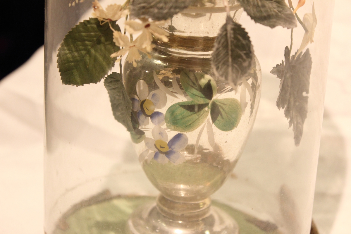 En "Glassmonter" med en blomsteroppsats i vase inne i. Blomstene ser ut som silkeblomster. Den har vært lukket av en ring i tre.