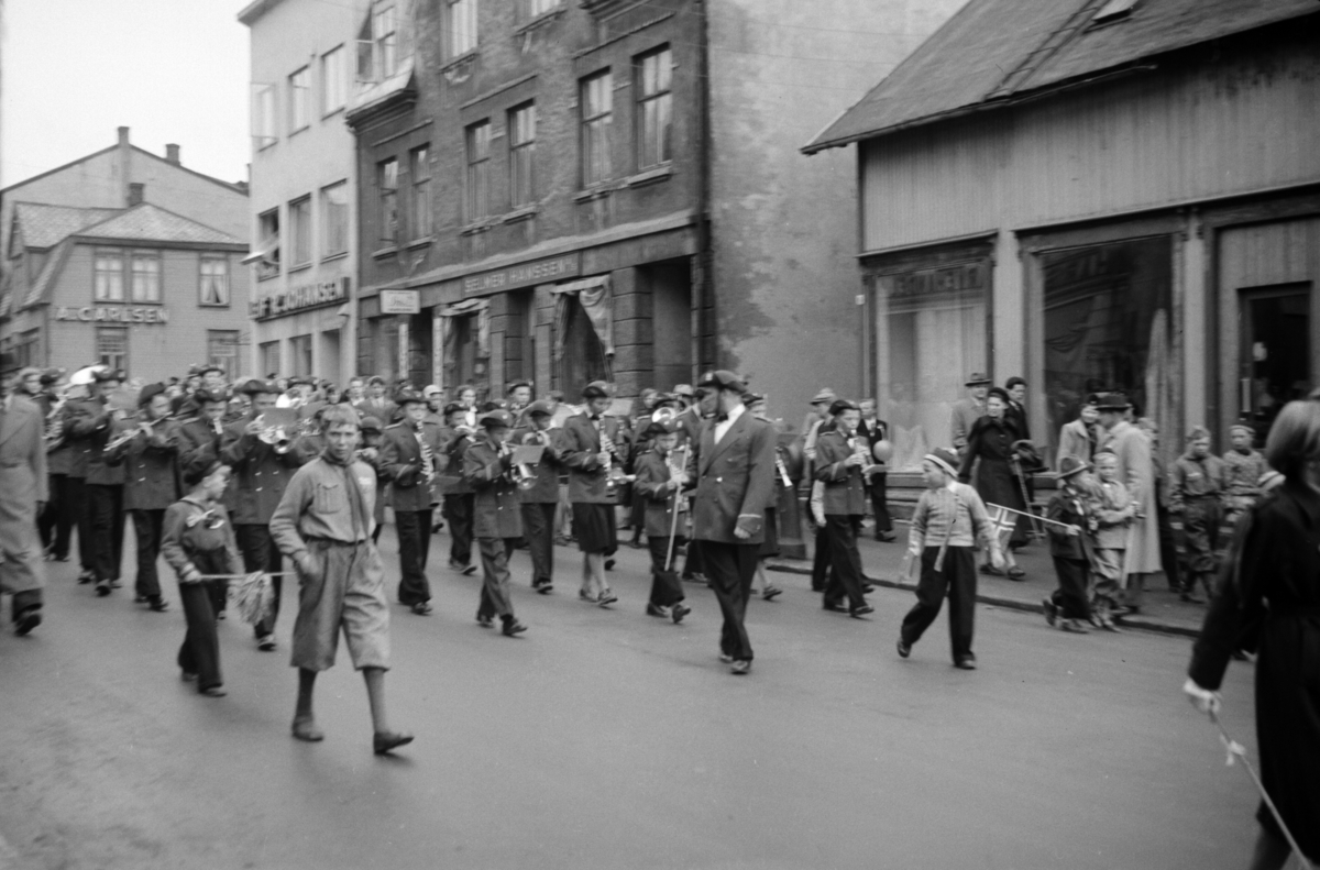 Skolekorps marsjerer ned Strandgata. I forgrunnen et par smågutter, samt en speider, i bakgrunnen flere butikkfasader.