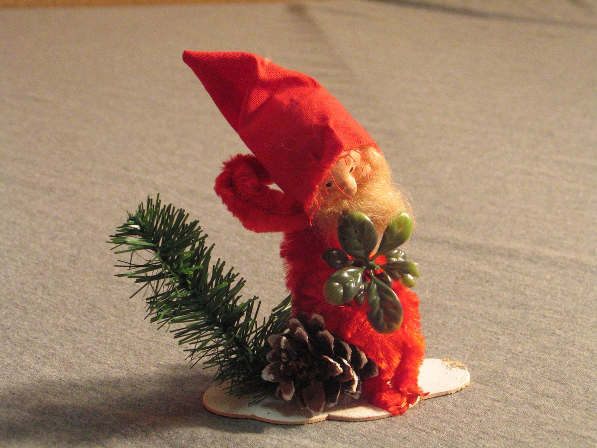 Julenisse av piperensar, sit på ei furukongle med snøglitter. Nissehovudet er av pappmache, måla ansikt, langt skjegg. Nissehue av stoff. Plastimitasjonar av granbar og tytebærlyng. 
Figuren er limt på ei papplate.