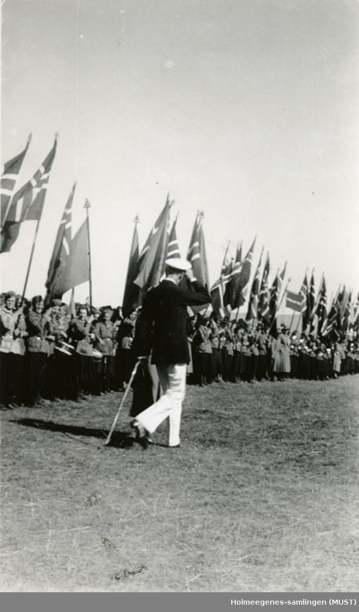 Kong Haakon VII og dronning Maud foran speidere med store norske flagg på speiderleir i 1935. Håndskrevet på baksiden av fotografiet: "Speiderne paraderer for kongen".