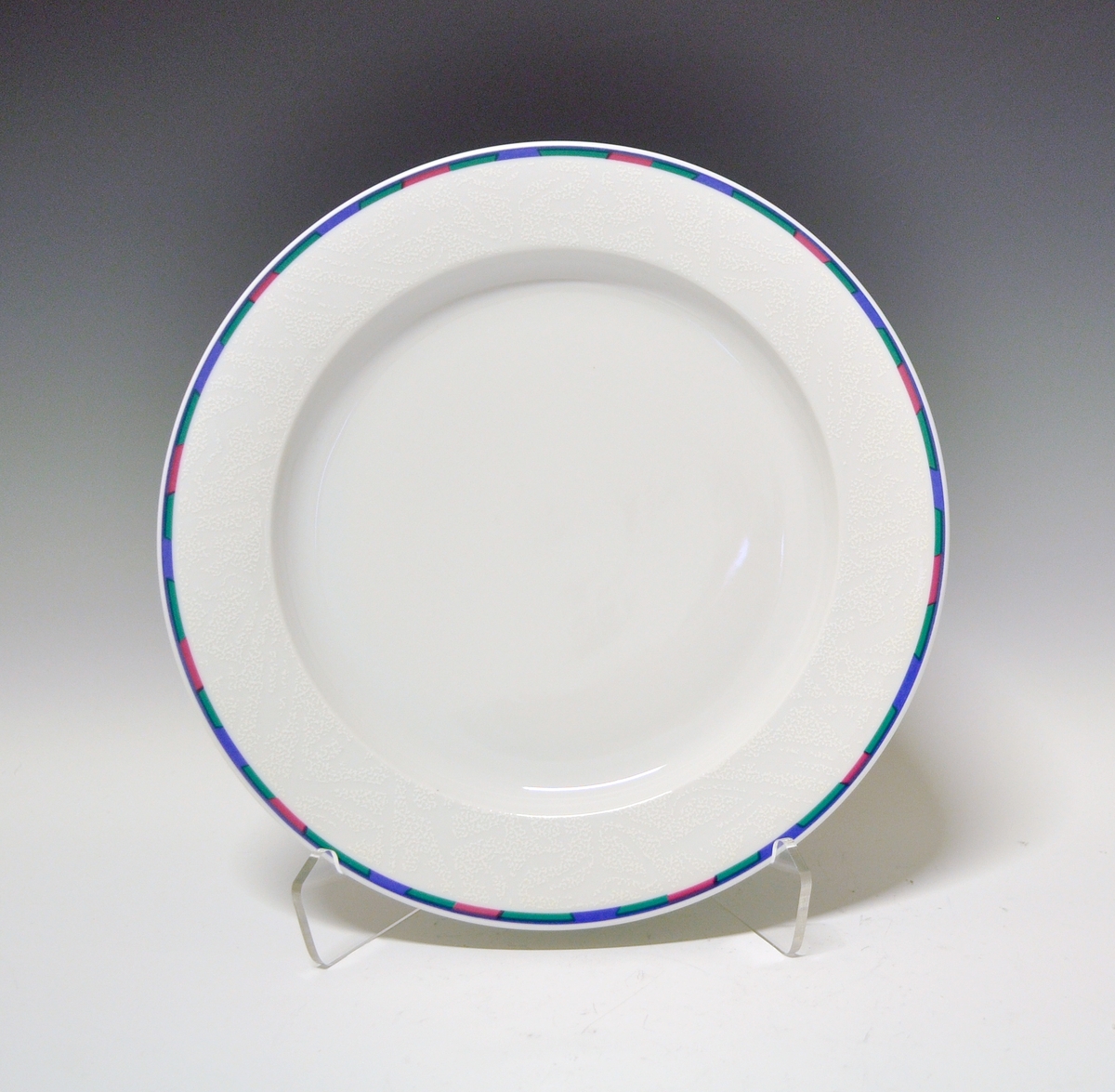 Middagstallerken av porselen med hvit glasur. Dekorert med rand ytterst på fanen i blått, grønt og rosa.
Modell: Saturn av Grete Rønning.