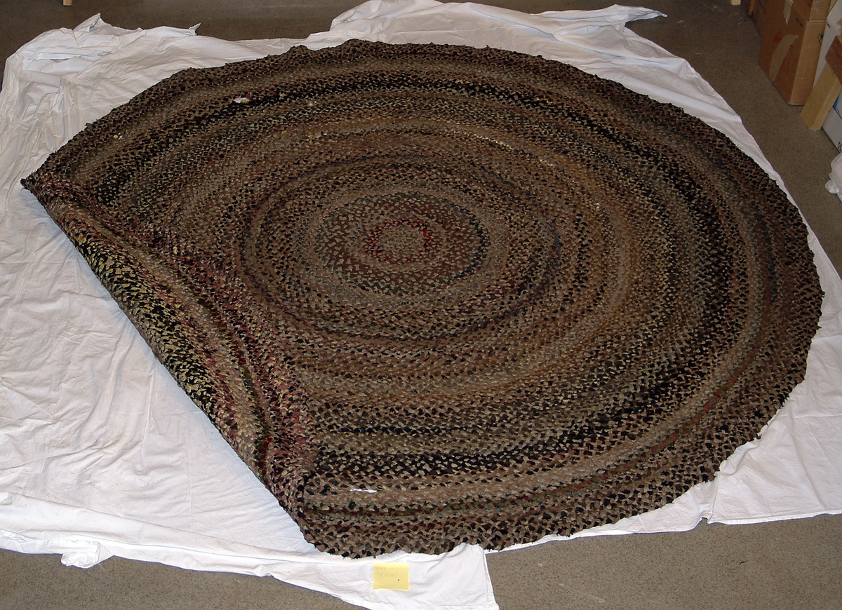 Rundt gulvteppe sammensydd av flettet tekstil. Flettingen består av 3 ulike tekstilremser som er skjøtet/sammensydd i endene. Fletteremsene er sydd sammen i en spiral med hvit bomullstråd på baksiden.