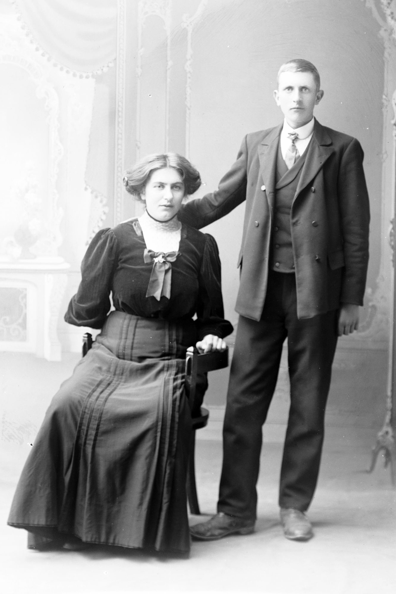 Studioportrett av en mann som står ved siden av en sittende kvinne.