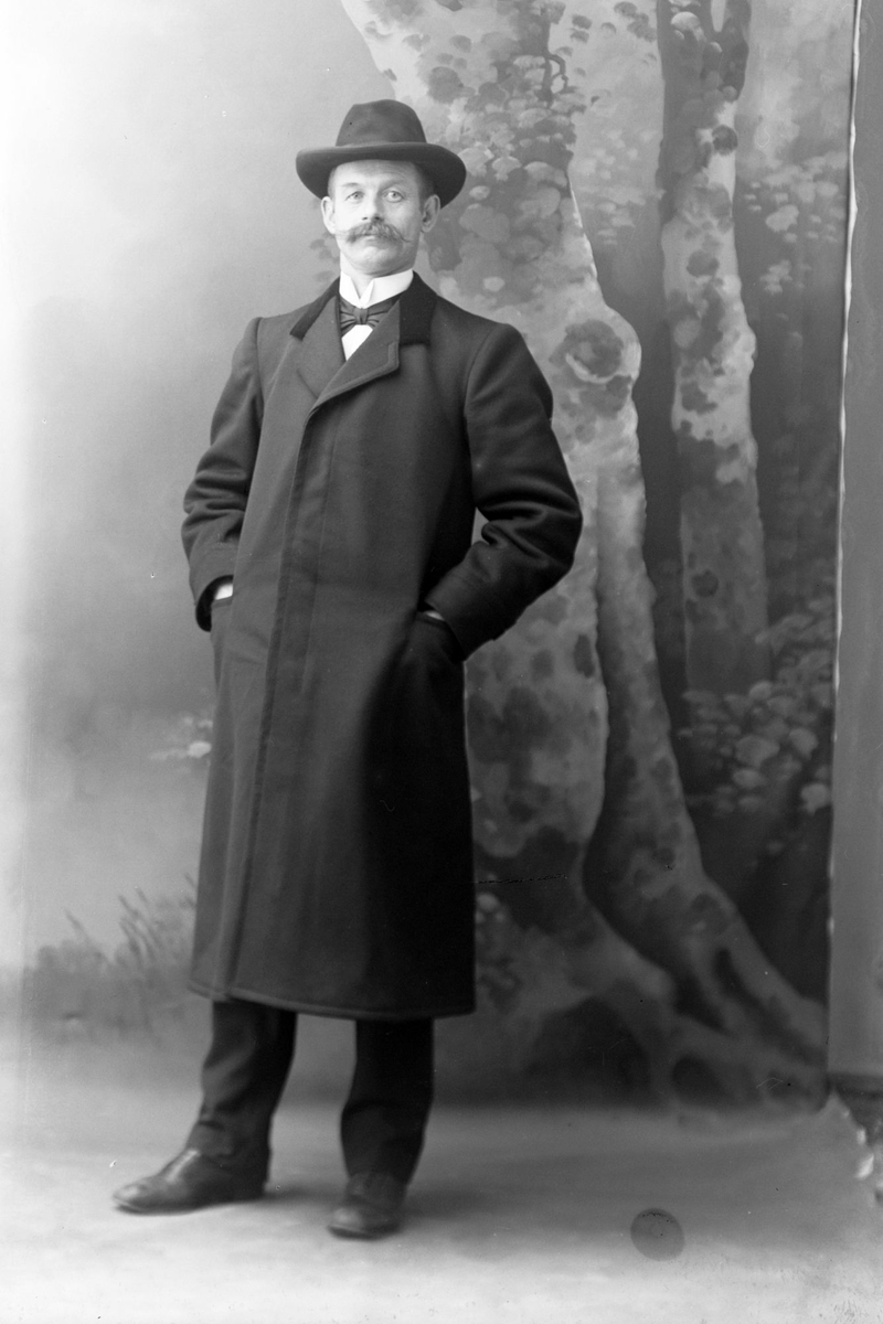 Studioportrett av en mann med hatt og lang frakk.