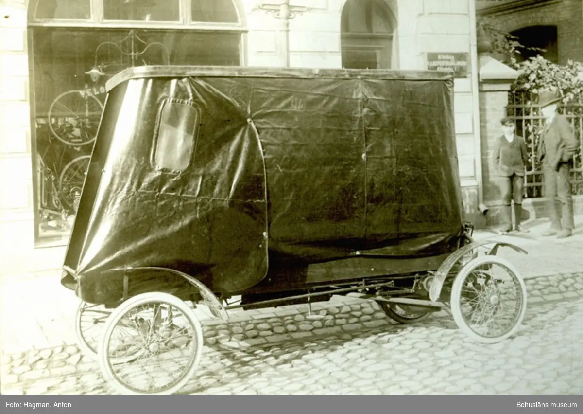 Bil med kapell parkerad på en kullerstensgata. Cykelaffär med text på glasrutan "Velocipedförsäljnings Aktiebolaget"