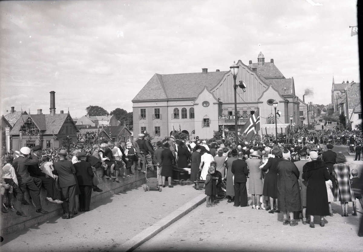 Stor folkemengde foran Rådhuset venter på kongens ankomst. Fotografert nordover Skåregt. Festiviteten midt på bildet. Fra kong Haakon VIIs besøk i Haugesund, den 22. august 1945.