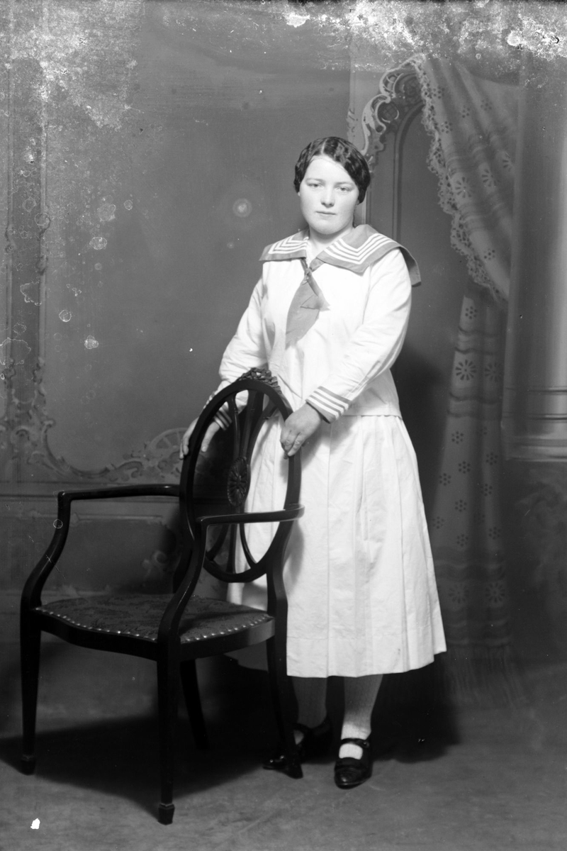 Studioportrett av en kvinne stående bak en stol.