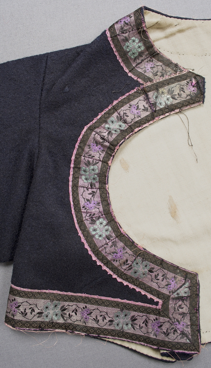 Mörkblå vadmalströja garnerad med band. Tröjan har en rund öppning fram på bröstet och skört bak. Runt halsringning och öppningen fram, längs nederkanten på framstycken och ärmar är tröjan dekorerad med ett 35 mm brett sidenband, bandet är mönstrat i lila, turkos, svart och rosa. Nederkanten på ärmen är dessutom dekorerad med ett mönstrat band i rosa och grönt silke på svart sammetsbotten. Bitar i form av hjärtan och en tulpan av ett blommönstrat band i starkt rosa och grönt är påsydda vid skörtet bak, detta ser ut att vara ett senare tillägg. Framtill knäpps tröjan på tre ställen med en hyska och hake.Ärmsprundet är 85 mm långt och har knäppts med en knapp; knapp saknas, tränsat knapphål. Axelbredd: 115 mm. Ärmlängd: 560 mm. Ärmen är sydd i ett stycke.Bakstycket har söm mitt bak. Tröjan är helfodrad med handvävt, tuskaftat halvblekt linne. Foder och band är påsydda för hand. Märkt på insidan av ena framstycket med en påsydd tyglapp med texten: "Landstinget N° 114 a".
